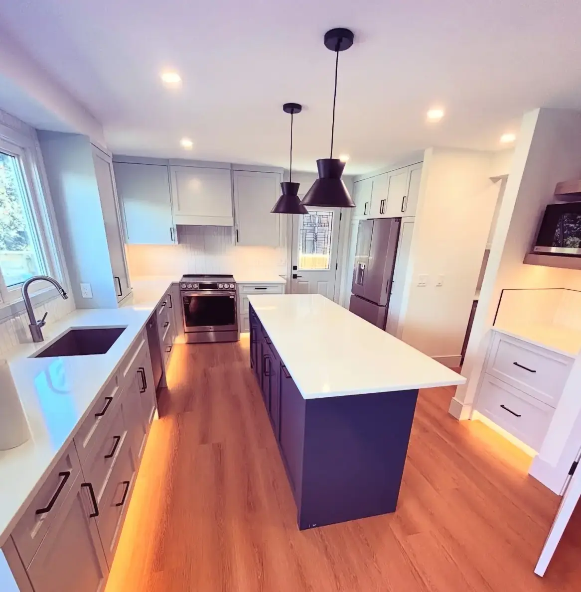 A beautifully renovated modern kitchen featuring light grey cabinets, a dark blue island, and strategically placed warm under-cabinet and toe-kick LED lighting, showcasing expert electrical installation work by The Urban Electrician